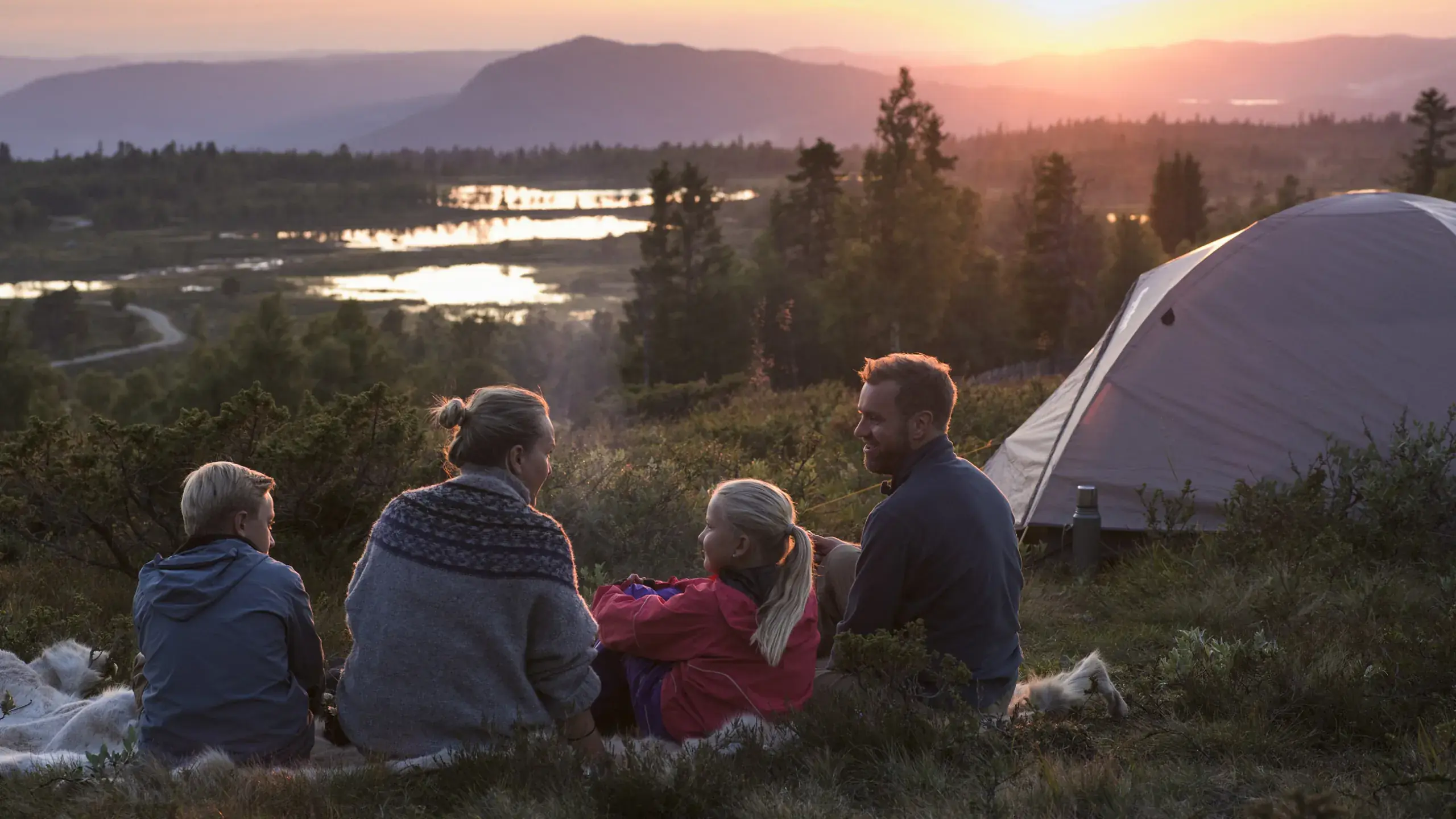 Folk sitter ved et telt og ser utover fjellvann i kveldssol
