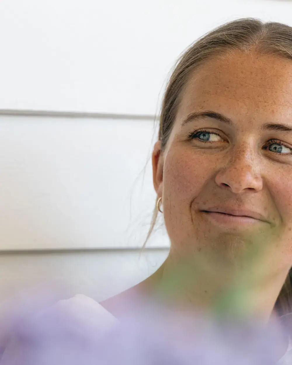 Nærbilde av Anette som smiler og ser til siden, foran en hvit trevegg, med lilla blomster uskarpt i forgrunnen.
