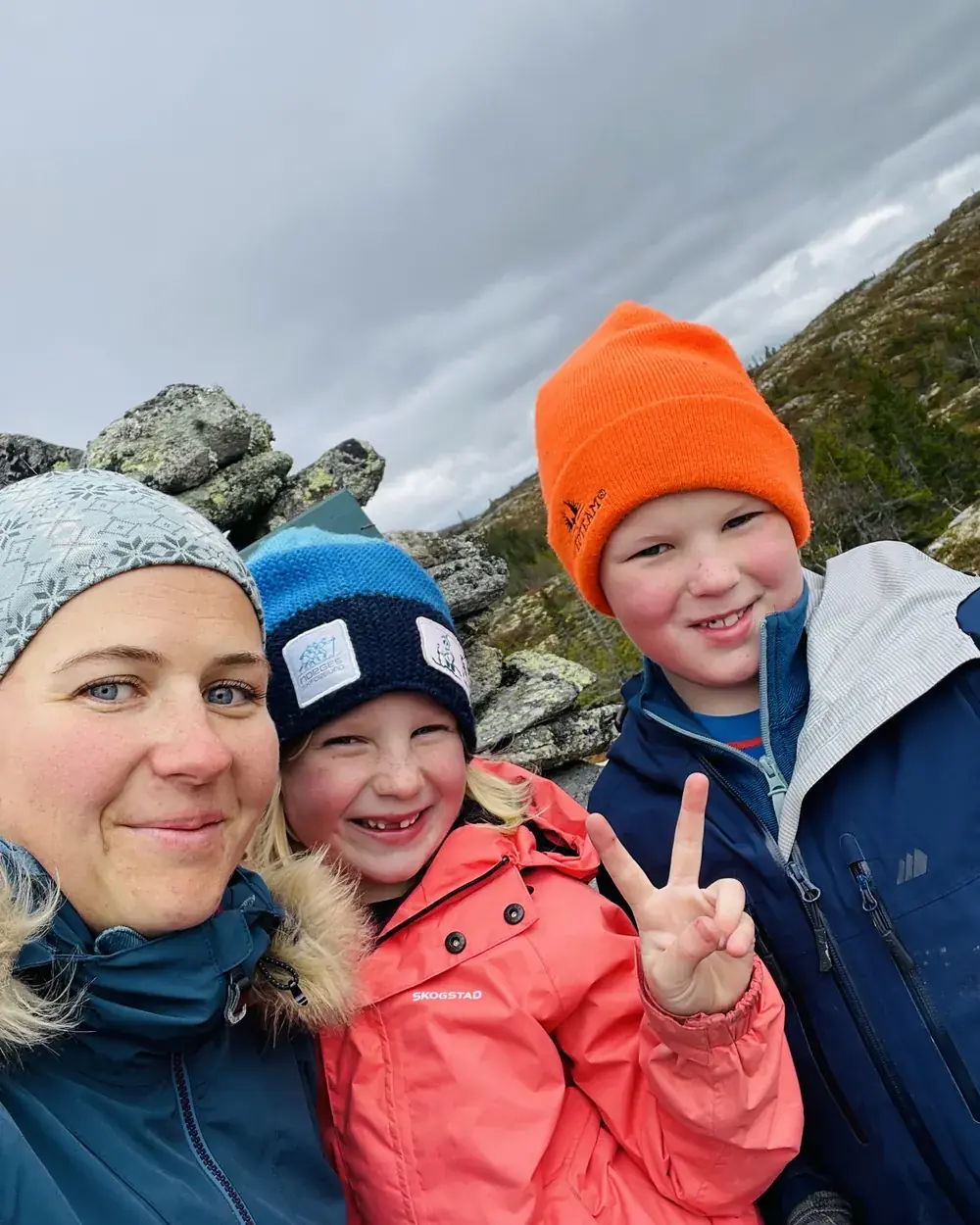 Anette i lue og vinterjakke tar en selfie med to smilende barn i strikkeluer på en steinete fjelltopp under grå himmel.