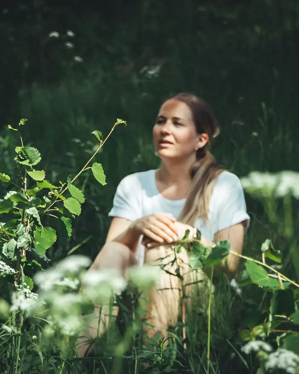 Anette sitter i høyt gress blant hvite skjermplanter og grønne blader, og ser ut til siden. Planter i forgrunnen er uskarpe.
