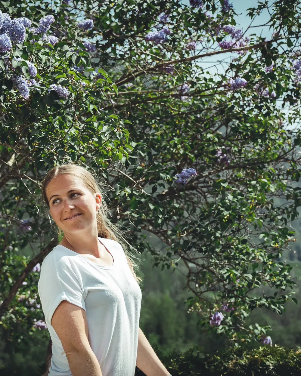 Anette i hvit t-skjorte smiler foran en blomstrende syrinbusk, med skogkledde fjellsider og blå himmel i bakgrunnen.
