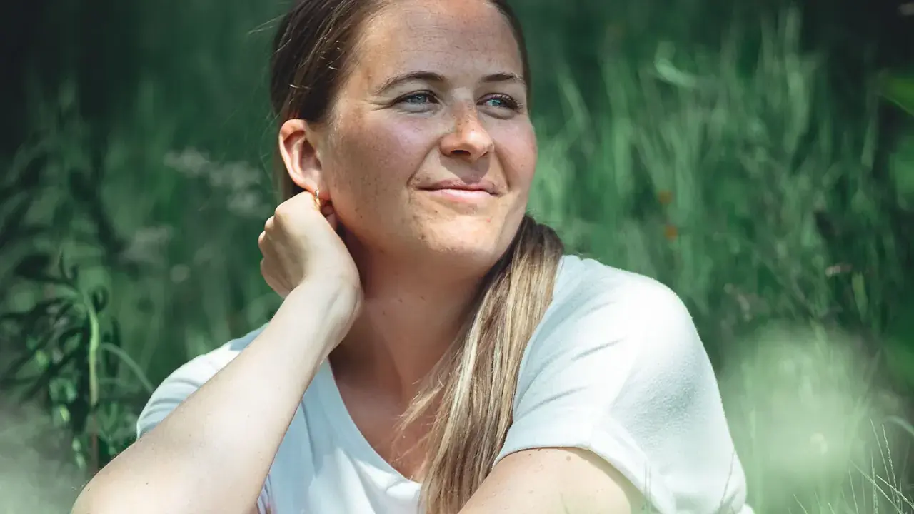 Anette sitter i høyt, grønt gress med hånda mot nakken og smiler, kledd i hvit t-skjorte.