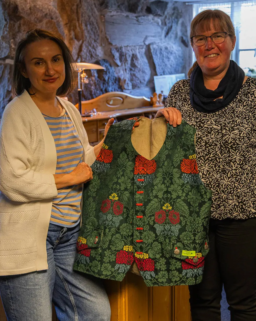 To kvinner holder opp en grønn vest i mønstret stoff med store røde blomster og en rekke røde knapper, foran en steinvegg.