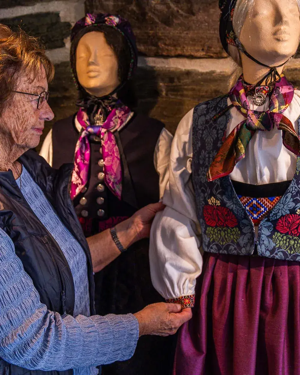 Gunhild Aasen retter på ermet til en bunad med blå, blomsterbrodert vest og rødt skjørt, blant flere bunader på utstillingsdukker.