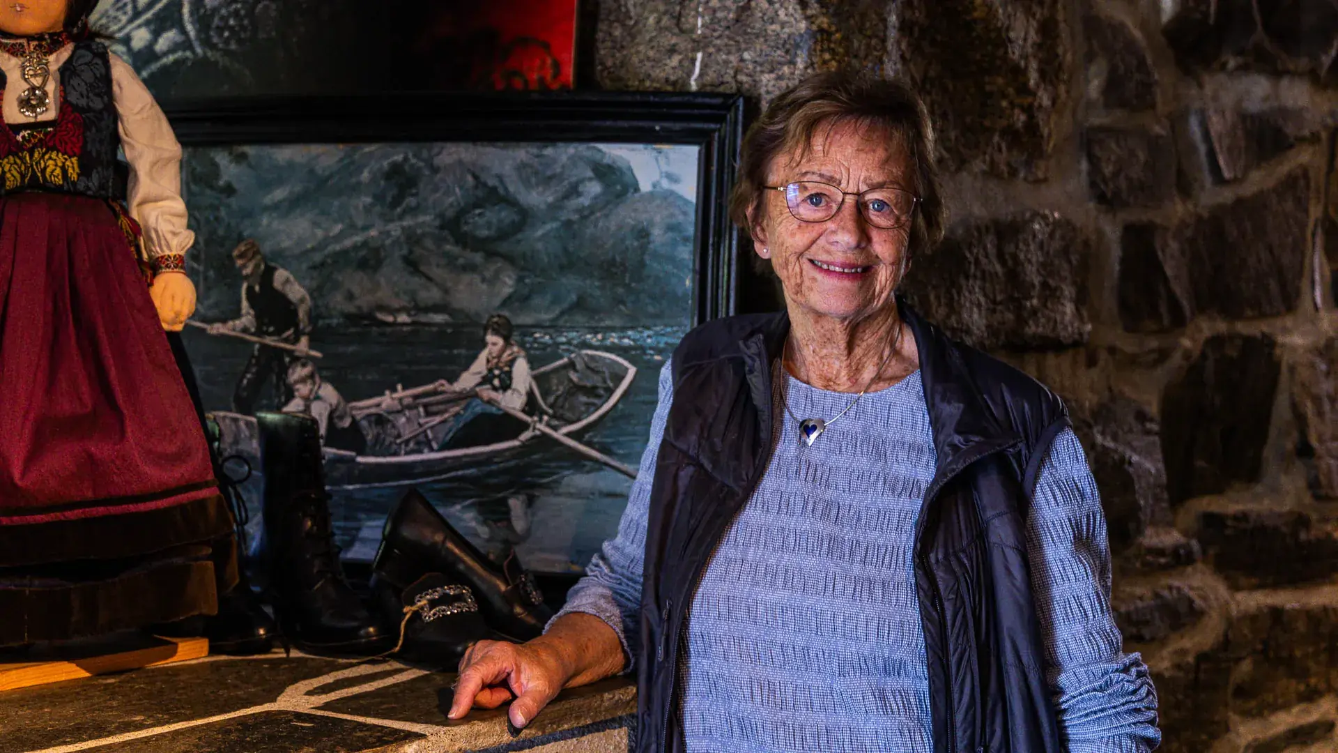 Gunhild Aasen smiler foran en steinvegg, med et maleri av folk i en robåt og en dukke i bunad ved siden av seg.