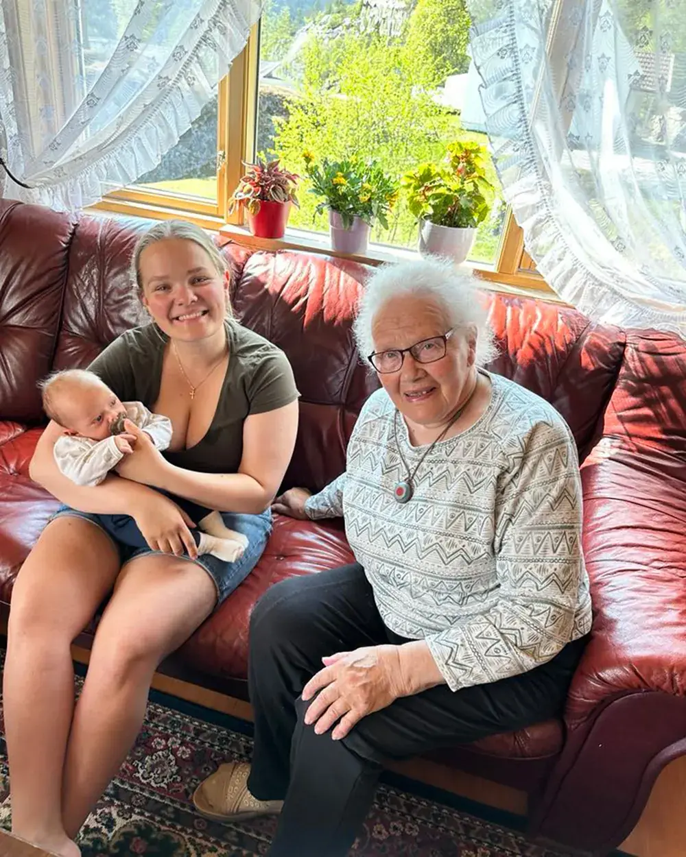 Ida sitter i en rød skinnsofa med en liten baby i armene, ved siden av en eldre kvinne med hvitt hår, foran et vindu med blomster i karmen.