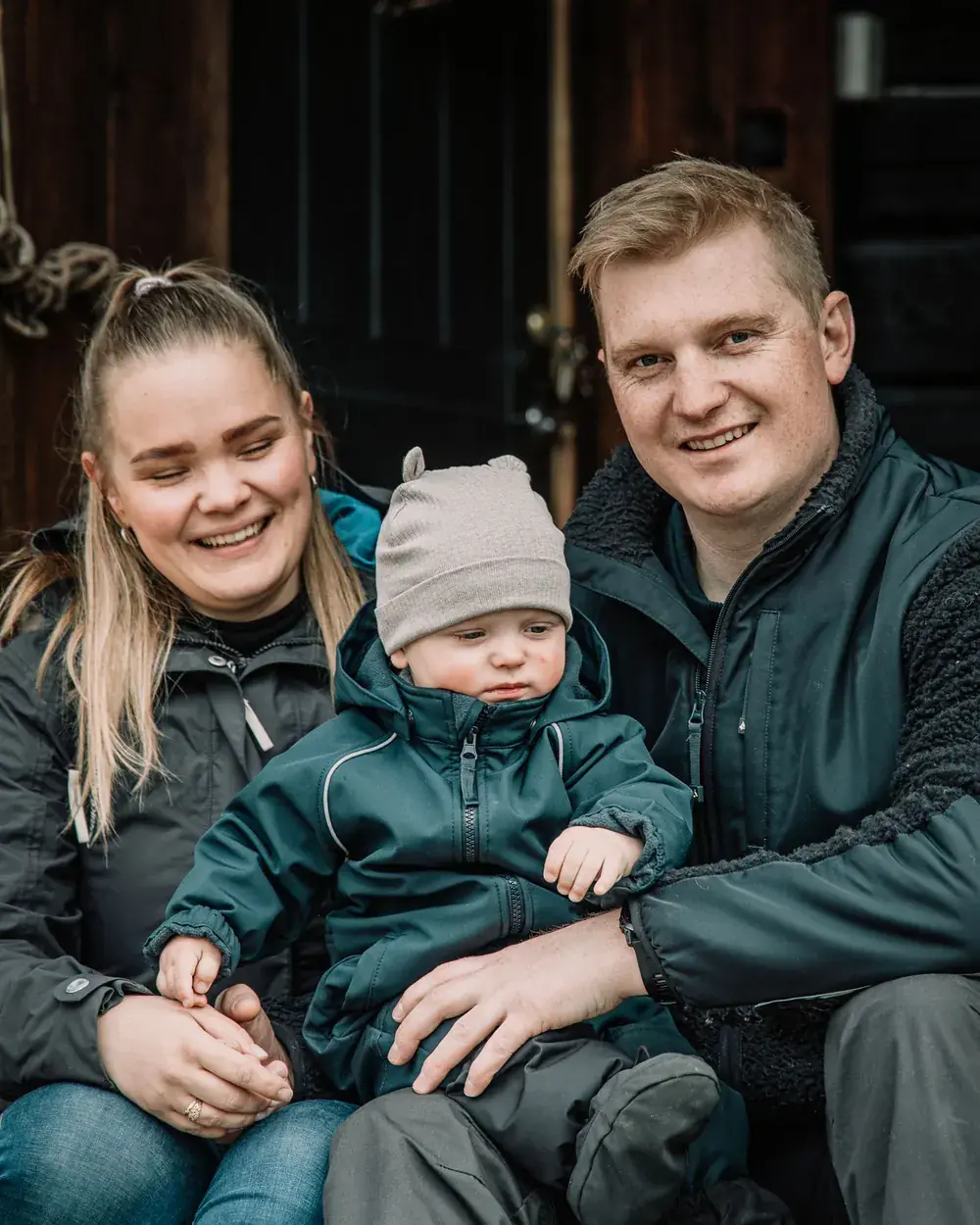 Ida og samboeren sitter smilende på en trapp foran en mørk tredør, med en liten gutt i grå lue og grønn dress på fanget.