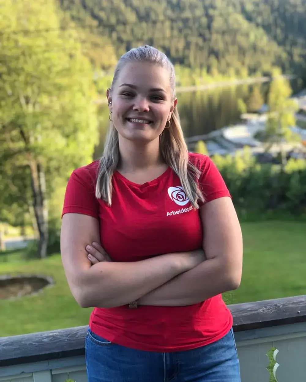 Ida står med armene i kors på en veranda i rød t-skjorte med Arbeiderpartiets logo, med grønn skogli og vann i dalen bak seg.