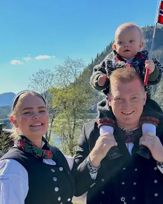 Ida og samboeren i bunad en solfylt dag, med sønnen på skuldrene og et norsk flagg i hånden, fjell og granskog i bakgrunnen.