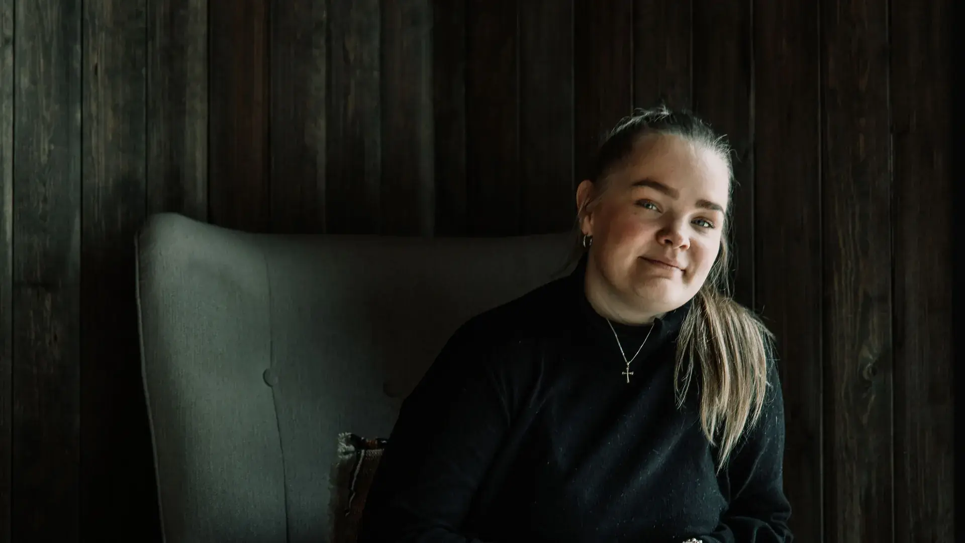Ida Bjerke sitter i en grå lenestol foran en mørk trevegg, i svart genser og med et lite kors i kjede rundt halsen.