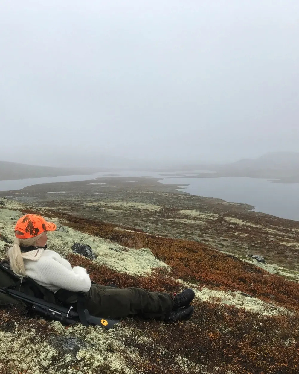 Liv-Astrid i oransje caps og hvit genser hviler mot sekken og rifla i høstfarget lyng, med utsikt over vann og fjellvidde i tåke.