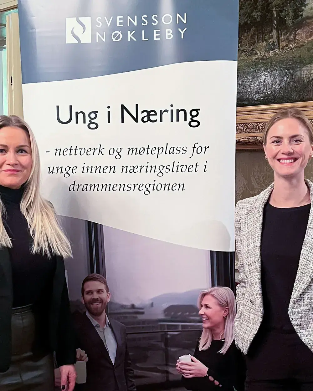 Liv-Astrid, til venstre, og en annen kvinne står foran et banner for Svensson Nøkleby og Ung i Næring i et rom med gullrammede malerier.