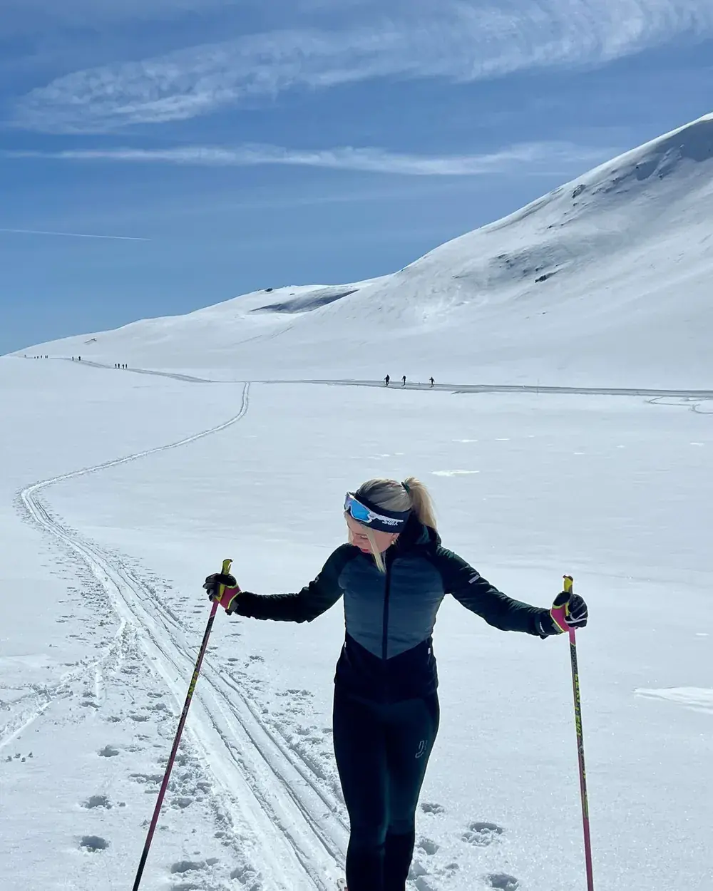 Liv-Astrid står med skistaver i et oppkjørt skispor på en snødekt fjellvidde under blå himmel, med flere skiløpere langt bak i løypa.