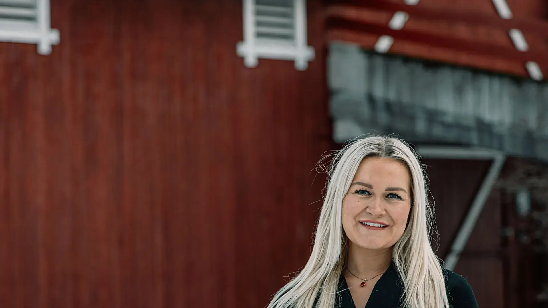 Liv-Astrid Rustgaard Bjerke med langt, lyst hår og svart kåpe smiler mot kamera foran en rød trevegg med hvite luker.
