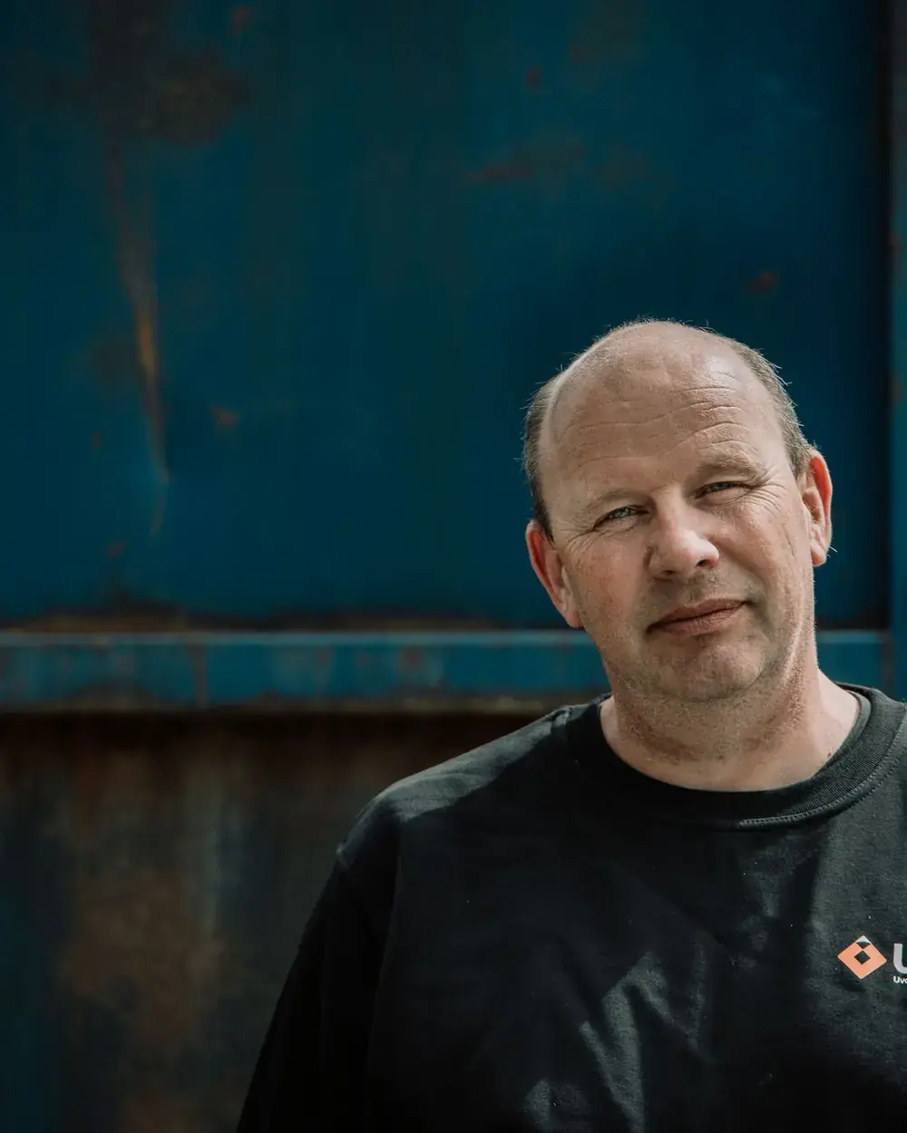 Ole Reidar i svart genser med UMF-logo står foran en blå, rustflekket stålcontainer og ser rolig inn i kamera.