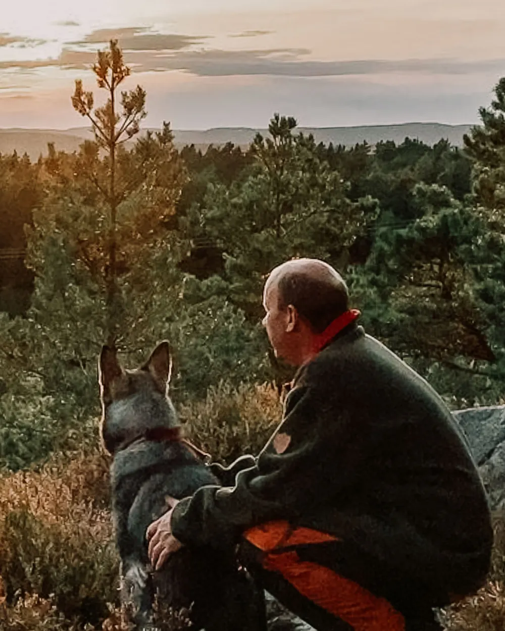 Ole Reidar sitter på huk i lyngen med armen rundt en grå hund. Begge ser utover skogkledde åser mens sola går ned.