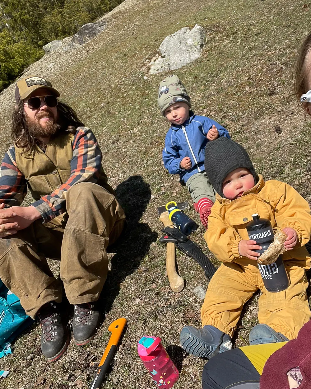 Familien hviler i en bratt, solfylt li. Sigmund sitter med solbriller og caps, de to barna sitter i gresset ved siden av ei øks, og Mari smiler inn i kameraet i forgrunnen.