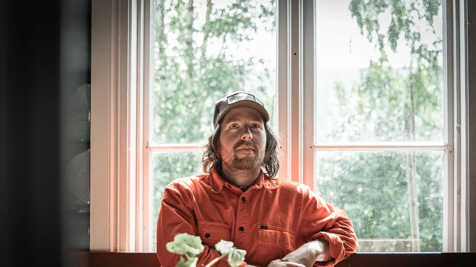 Sigmund i oransje skjorte og caps lener seg mot en benk foran et stort sprossevindu med grønne bjørker utenfor. En pelargonia står i forgrunnen.