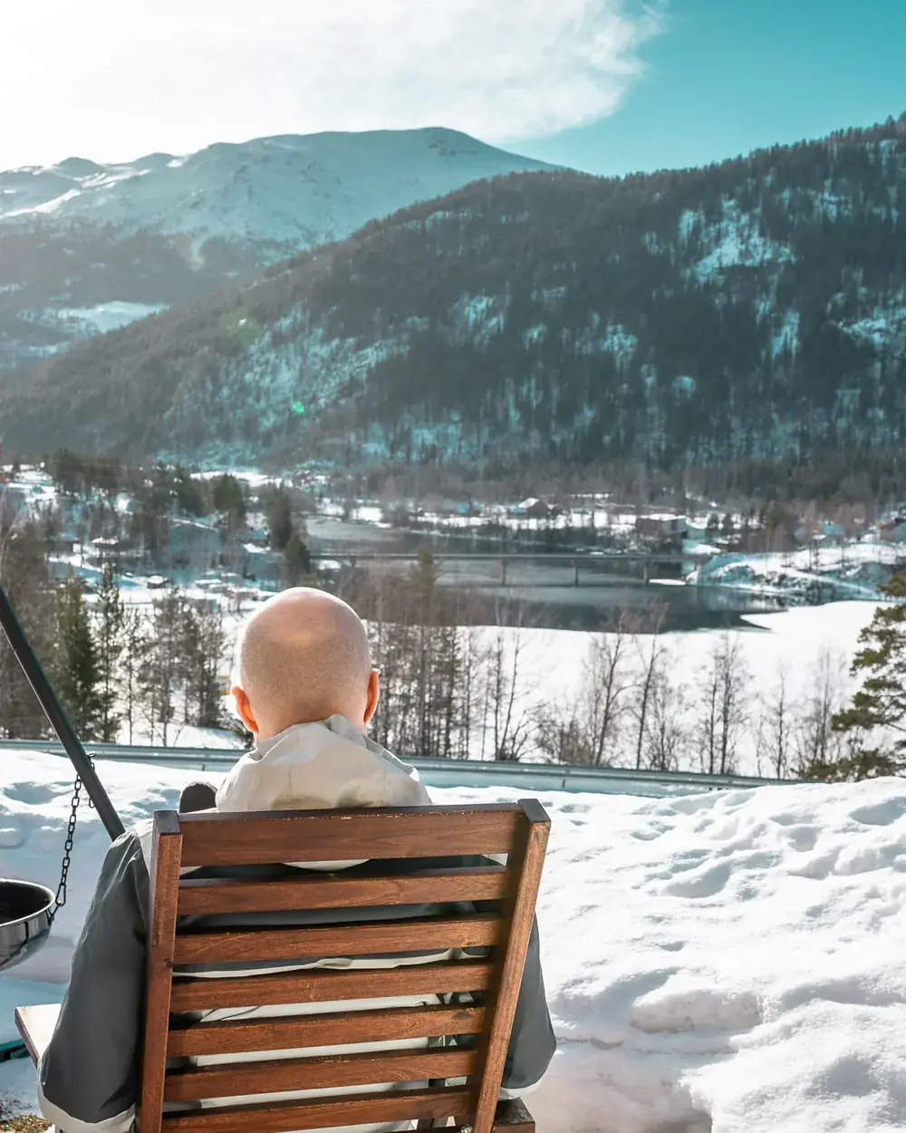 Sveinung sitter med ryggen til i en trestol i snøen og ser utover en dal med elv, bro, hus og en hvit kirke, med snødekte fjell i bakgrunnen.