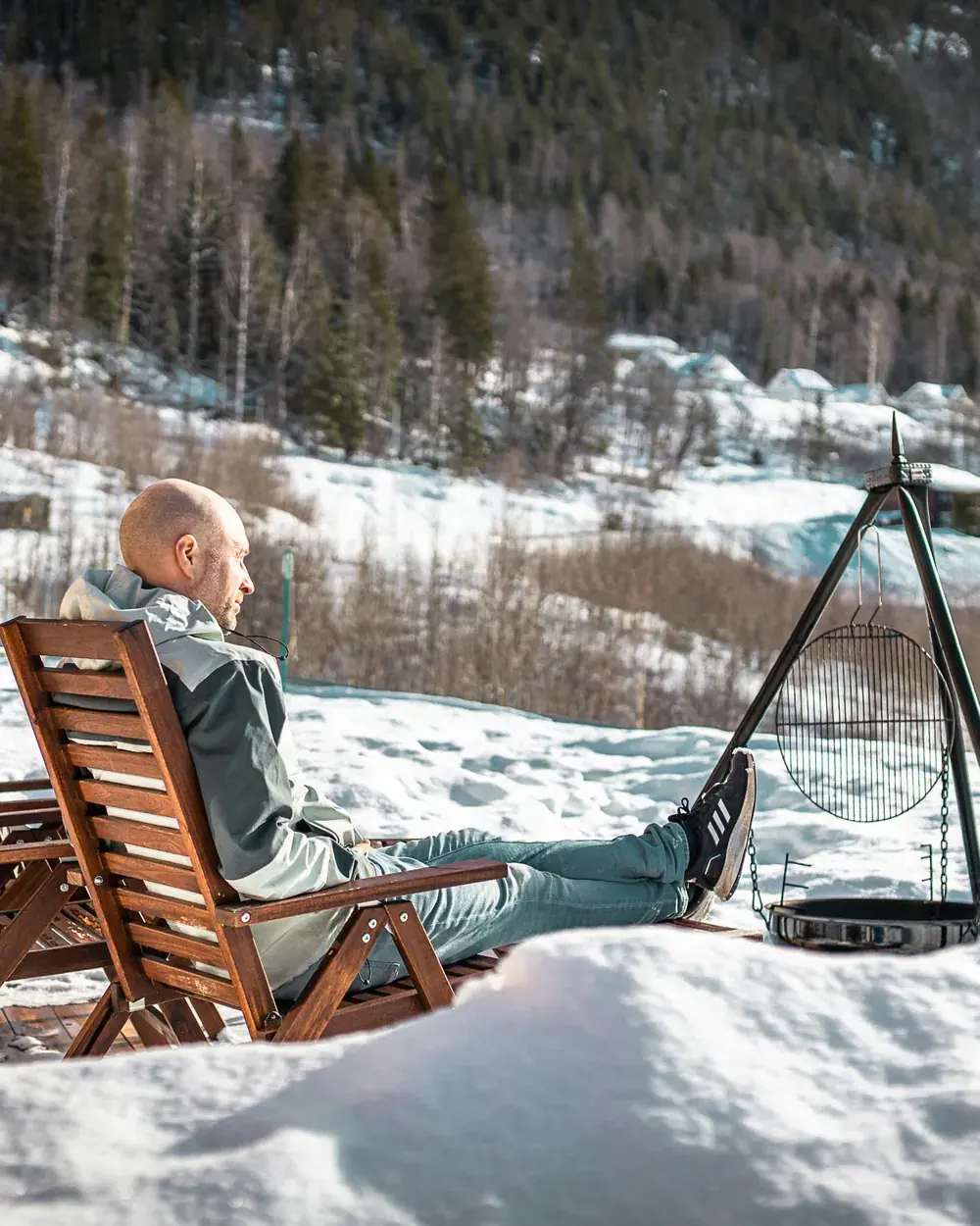 Sveinung lener seg tilbake i en trestol i snøen med beina høyt, ved siden av et bålpanne-stativ med grillrist, med skogkledd li og hus i bakgrunnen.