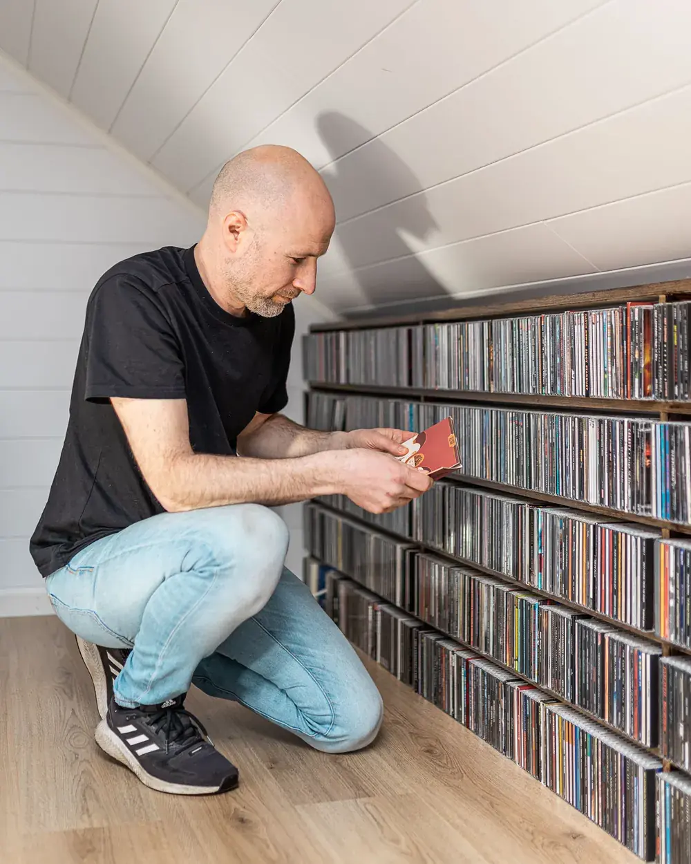 Sveinung sitter på huk i et rom med skråtak og blar i et CD-cover foran en lang, lav hylle full av CD-er.