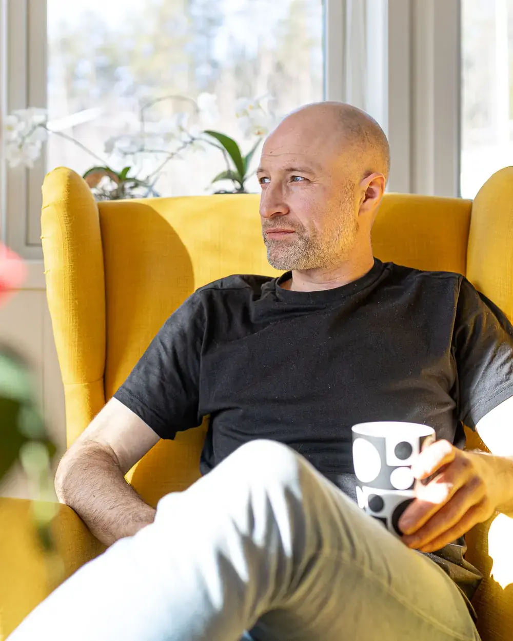 Sveinung sitter i en gul ørelappstol i sollyset og ser ut av vinduet med en kaffekopp i hånda, med blomster i vinduskarmen og et strikket teppe over stolen ved siden av.