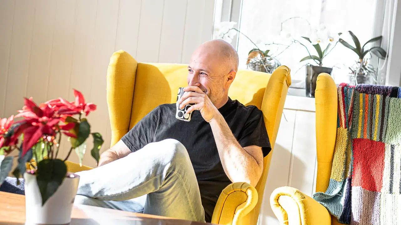 Sveinung Dahl i svart t-skjorte sitter smilende i en gul ørelappstol med en mønstret kaffekopp, med sollys, orkideer i vinduet og en julestjerne i forgrunnen.