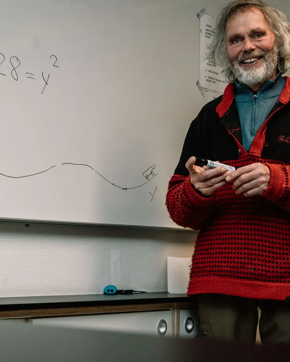 Tov Sandnæs smiler med en tusj i hendene foran en tavle med en matematisk likning og en tegnet kurve.
