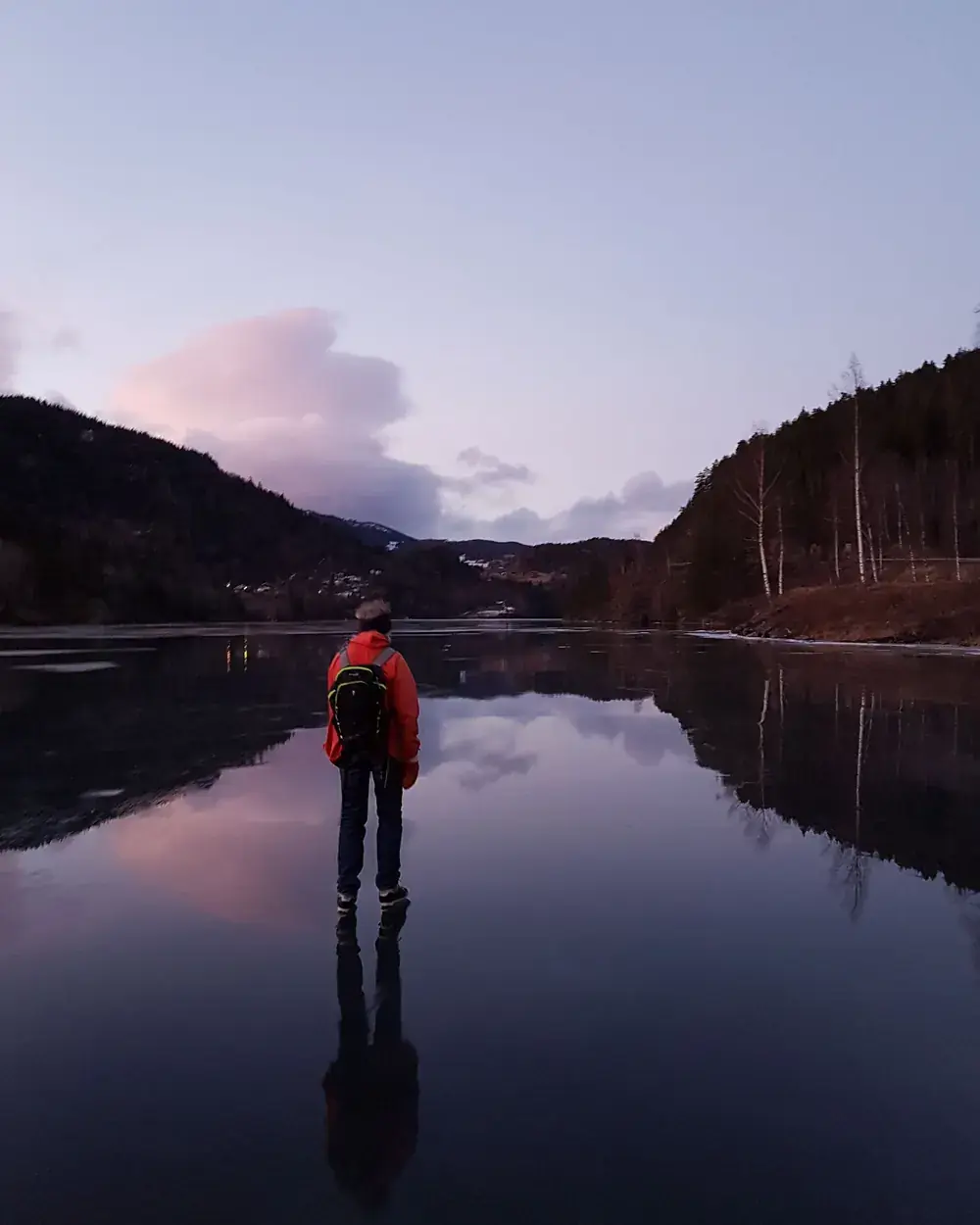 En person i rød jakke og ryggsekk står på blank is på fjorden i skumringen, med åser og rosa skyer speilet i isen.