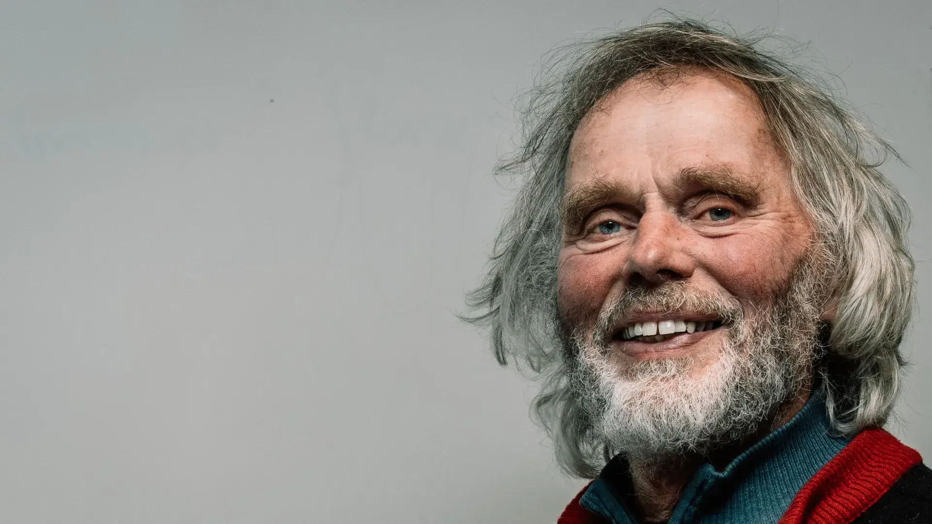 Portrett av Tov Sandnæs som smiler, med langt grått hår og grått skjegg, i blå genser og mørk jakke med rød krage.